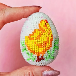 以下が含まれることがあります： 黄色いひよこのデザインが施された白いビーズのイースターエッグ。ひよこは緑の葉とピンクの花に囲まれています