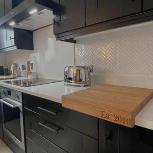The Modern Kitchen Display: A personalized solid oak chopping board with "Est. 2010" laser-engraved on the front stability lip. It is shown protecting a white worktop in a contemporary kitchen with black cabinetry and a white herringbone tile backsplash.