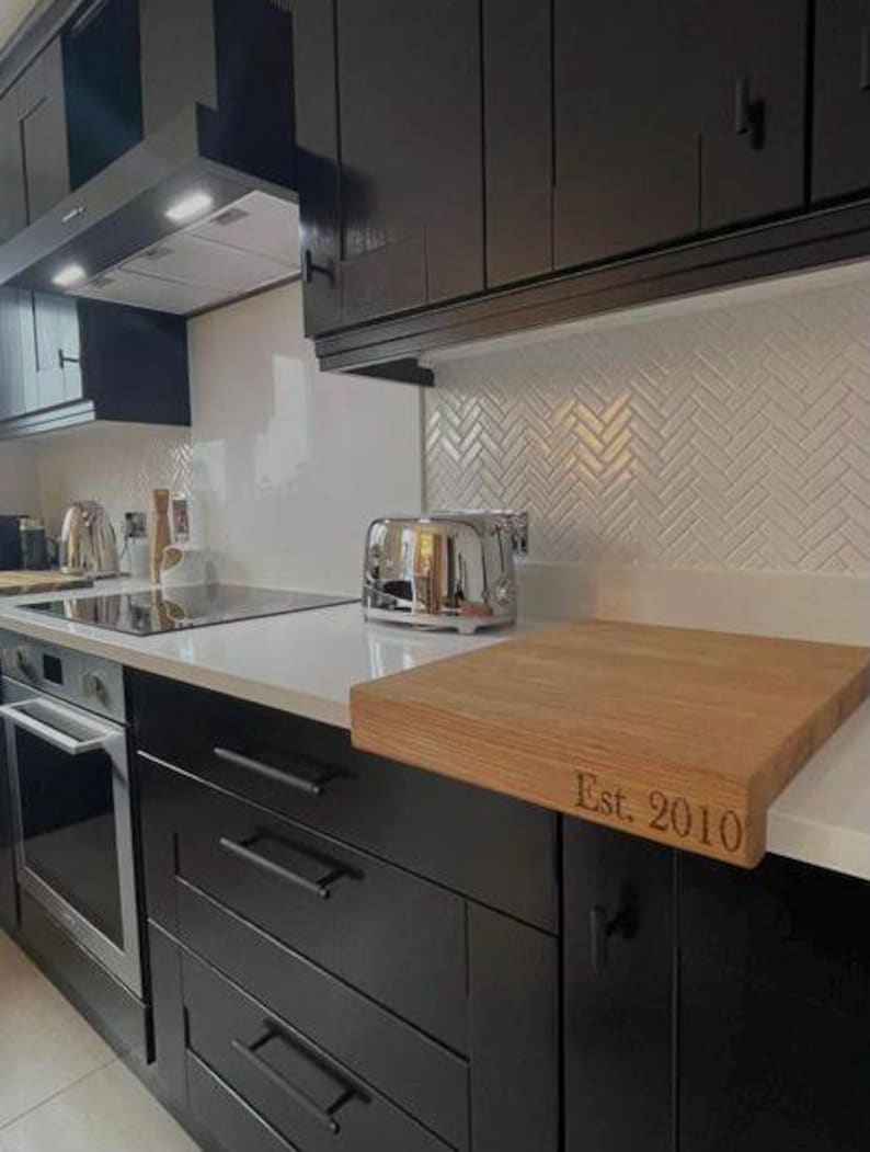 The Modern Kitchen Display: A personalized solid oak chopping board with "Est. 2010" laser-engraved on the front stability lip. It is shown protecting a white worktop in a contemporary kitchen with black cabinetry and a white herringbone tile backsplash.