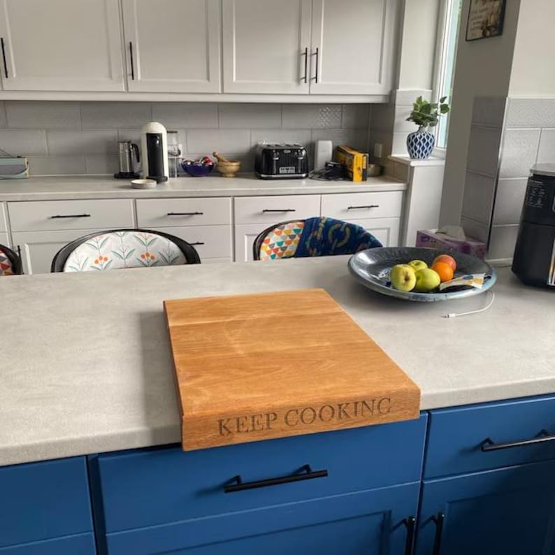 Solid oak worktop saver chopping board with counter lip overhang, laser engraved with "KEEP COOKING" on the front edge. Displayed on a grey kitchen island with blue cabinets.
