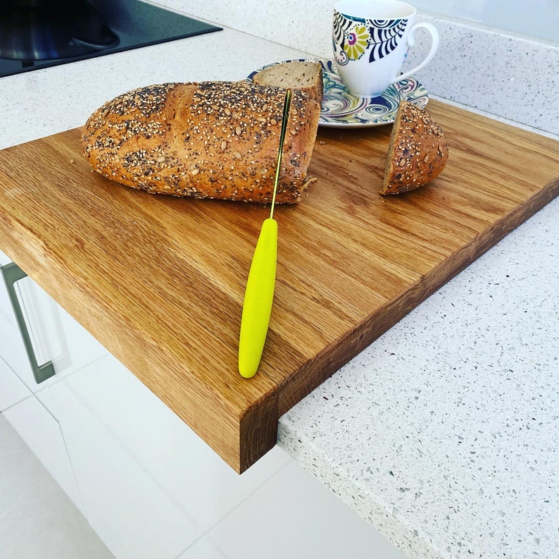 A large, thick solid oak chopping board with a unique "lip" design that hooks over the edge of a white quartz kitchen counter. On top sits a crusty seeded loaf of bread being sliced by a yellow-handled knife, a patterned tea cup, and a slice of bread on a matching saucer. The oak has a rich, warm grain and a smooth finish. The board is shown in a bright, modern kitchen setting, demonstrating its sturdy fit against the countertop edge.