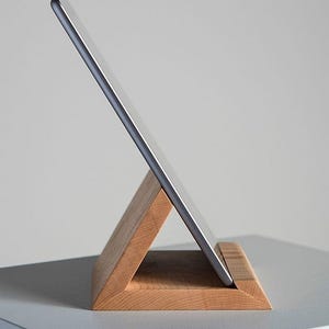 May include: A wooden tablet stand with a triangular base. The stand is made of light brown wood and has a smooth finish. A silver tablet is resting on the stand.
