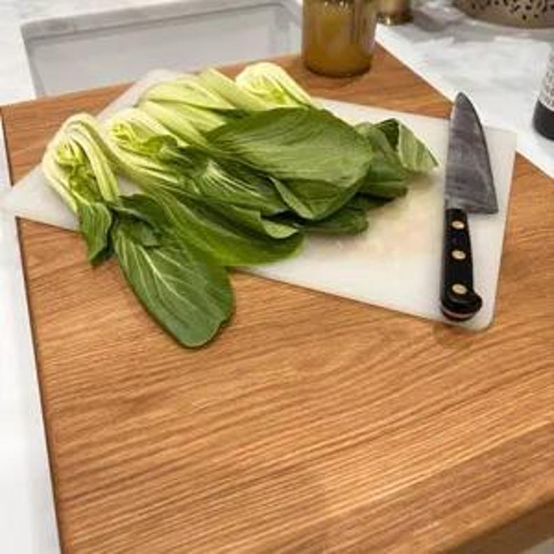Large solid oak chopping board and worktop saver shown in action with fresh pak choi and a chef's knife on a thin cutting mat resting on top. Handmade wooden counter lip board in a kitchen.