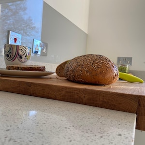 Lifestyle Scene (Bread & Coffee): A sliced seeded loaf of bread and a coffee cup resting on a large solid oak worktop protector, highlighting the natural wood grain and its secure placement on a light-colored worktop.