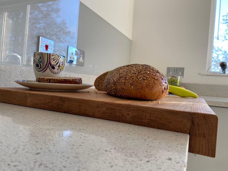 Lifestyle Scene (Bread & Coffee): A sliced seeded loaf of bread and a coffee cup resting on a large solid oak worktop protector, highlighting the natural wood grain and its secure placement on a light-colored worktop.