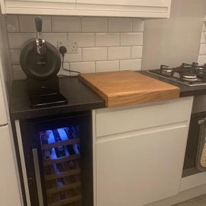 Solid oak worktop saver chopping board with counter lip, resting on a black kitchen counter next to a Krups beer dispenser and gas hob, above an under-counter wine fridge.