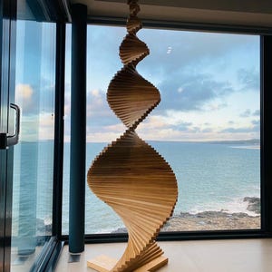 Op de afbeelding: Een houten sculptuur met een spiraalvormig ontwerp. De sculptuur is gemaakt van lichtbruin hout en heeft een ster aan de bovenkant. De basis heeft een kruisvorm. De sculptuur staat voor een groot raam met uitzicht op de zee.