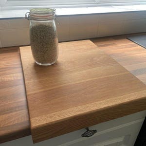 Solid oak worktop saver chopping board with counter lip overhang, featuring a glass storage jar of oats on top. Resting on a wood-grain kitchen countertop near a window.