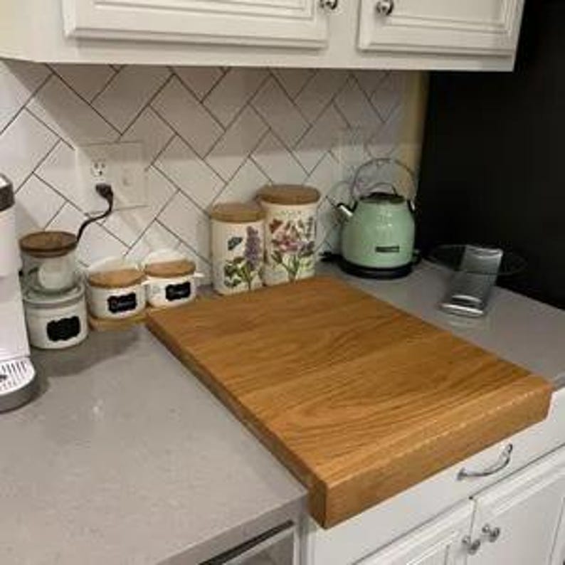 Solid oak worktop saver chopping board with counter lip, sitting flat on a grey kitchen countertop against a white herringbone tile backsplash. Large wooden counter-edge butcher block.