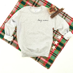 May include: A gray sweatshirt with the text "dog mom" embroidered on the front. The sweatshirt is lying on a red and green plaid blanket.