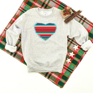 Op de afbeelding: Een grijs sweatshirt met een kleurrijk gestreept hartontwerp. Het hart is omrand met blauw en heeft een verweerde look. Het sweatshirt ligt op een rood-groen geruit deken.