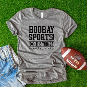 Op de afbeelding: Een grijs T-shirt met de tekst "HOORAY SPORTS! DO THE THINGS win the points" erop gedrukt. Het shirt ligt op een groene ondergrond met een bruine voetbal ernaast.