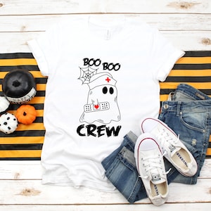 May include: White t-shirt with a Halloween-themed graphic. The design features a ghost wearing a nurse's cap and a bandage with a heart, with the words "BOO BOO CREW". The shirt is paired with blue jeans and white sneakers.