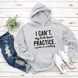 May include: A light gray hooded sweatshirt with the text "I CAN'T. my kids have PRACTICE. a game or something" in black. The sweatshirt is paired with blue jeans and white sneakers. A small green plant is also in the image.