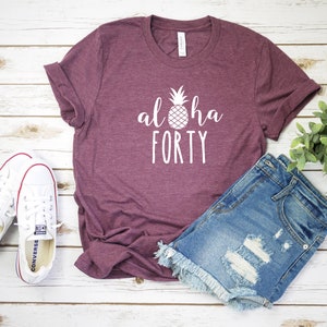 May include: A maroon t-shirt with the words "aloha FORTY" and a pineapple graphic in white. The shirt is paired with denim shorts and white canvas sneakers. The items are arranged on a light wood surface.