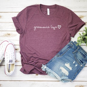 May include: Burgundy t-shirt with the text "grammie life" in white with a small heart next to it. The shirt is laying on a white wooden surface with a pair of white sneakers and a pair of blue denim shorts with frayed edges.