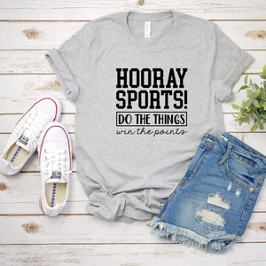 Op de afbeelding: Een grijs T-shirt met de tekst "HOORAY SPORTS! DO THE THINGS win the points" in zwart gedrukt. Het shirt ligt op een houten oppervlak met een paar witte sneakers en een blauwe spijkerbroek.