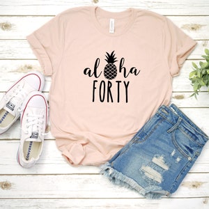 May include: A light pink t-shirt with the words "aloha FORTY" and a pineapple graphic in black. The shirt is paired with denim shorts and white sneakers with red accents.