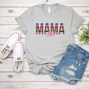 Op de afbeelding: Grijs T-shirt met de tekst "MAMA cita" in kleurrijke gestreepte letters. De letters zijn in een kleurrijk gestreept patroon dat doet denken aan een Mexicaanse deken. Het shirt ligt plat op een wit houten oppervlak met een paar witte canvas schoenen en een blauwe denim short.