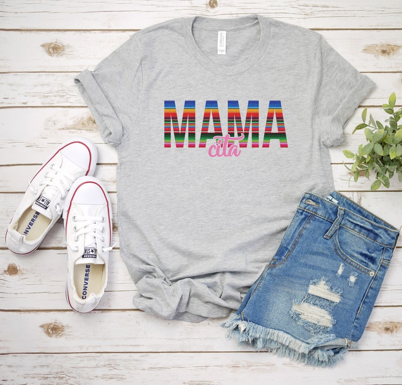 Op de afbeelding: Grijs T-shirt met de tekst "MAMA cita" in kleurrijke gestreepte letters. De letters zijn in een kleurrijk gestreept patroon dat doet denken aan een Mexicaanse deken. Het shirt ligt plat op een wit houten oppervlak met een paar witte canvas schoenen en een blauwe denim short.