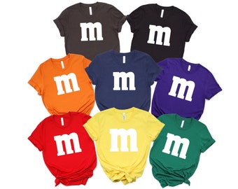 May include: Eight t-shirts in different colors with a white "M" printed on the front. The colors are orange, red, yellow, green, blue, purple, gray, and black.
