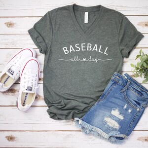 May include: A dark green V-neck t-shirt with the text "BASEBALL all day" in white lettering. The shirt is laid flat on a white wooden surface with a pair of white sneakers and a pair of blue denim shorts.