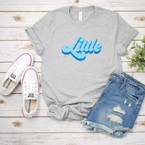 May include: A gray t-shirt with the word "Little" in a blue retro font. The shirt is laid out on a white wooden surface with a pair of white sneakers and a pair of blue denim shorts.