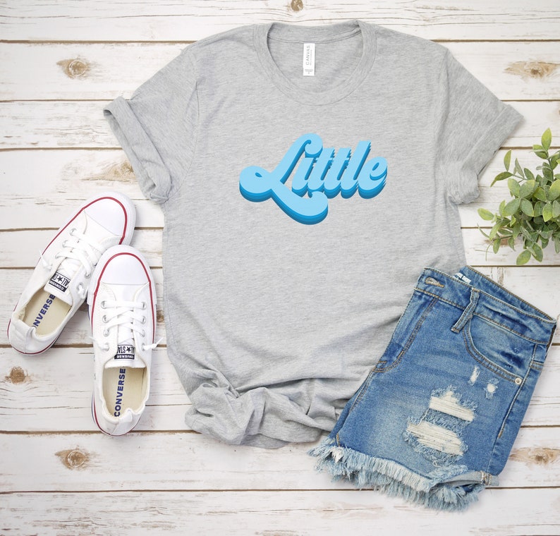 May include: A gray t-shirt with the word "Little" in a blue retro font. The shirt is laid out on a white wooden surface with a pair of white sneakers and a pair of blue denim shorts.