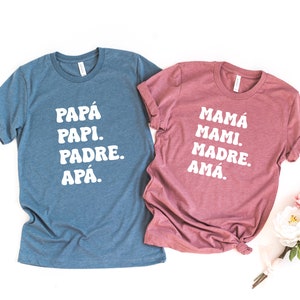 May include: Two t-shirts with Spanish words for "dad" and "mom". The blue shirt says "Papá Papi Padre Apá." The pink shirt says "Mamá Mami Madre Amá."