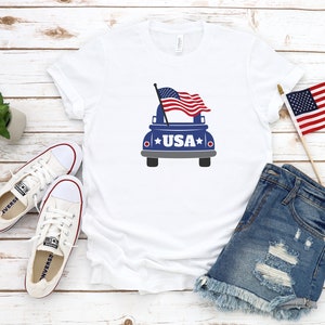 Op de afbeelding: Wit T-shirt met een blauwe pick-up truck die een Amerikaanse vlag draagt en het woord "USA" op de achterkant gedrukt. Het shirt ligt op een wit houten oppervlak met een paar witte canvas schoenen, een spijkerbroekje en een kleine Amerikaanse vlag.