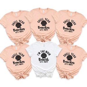 May include: Five peach-colored t-shirts with black text and a black seashell graphic. One white t-shirt with black text and a black seashell graphic. The text on the peach-colored shirts reads "Shell Yeah Beaches Let's Party!" The text on the white shirt reads "I'm the Main Beach Let's Party!"