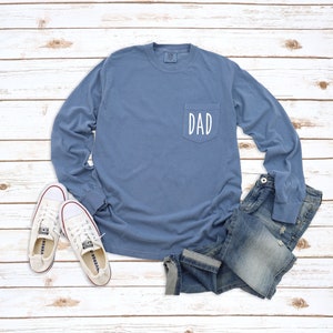 May include: A blue long-sleeved t-shirt with a pocket on the front. The pocket has the word "DAD" written on it in white.