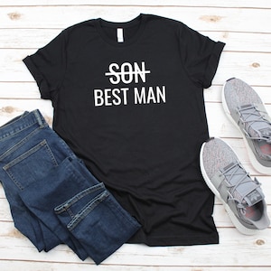 May include: Black t-shirt with white text that says "SON" with a line through it and "BEST MAN" underneath. The shirt is laid flat on a white wooden surface with a pair of gray sneakers and blue jeans.