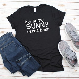 Pode incluir: Camiseta preta com texto branco que diz "some bunny needs beer" e um desenho de orelha de coelho de desenho animado.