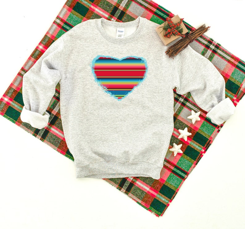 Op de afbeelding: Een grijs sweatshirt met een kleurrijk gestreept hartontwerp. Het hart is omrand met blauw en heeft een verweerde look. Het sweatshirt ligt op een rood-groen geruit deken.