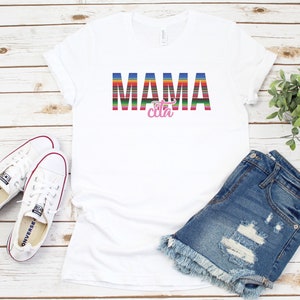 Op de afbeelding: Wit T-shirt met de tekst "MAMA cita" in kleurrijke gestreepte letters. Het woord "cita" is roze. Het shirt ligt plat op een witte houten achtergrond met een paar witte sneakers, een spijkerbroekje en een plant in een pot.