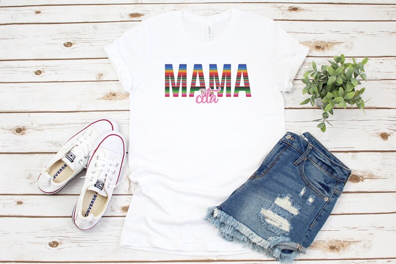 Op de afbeelding: Wit T-shirt met de tekst "MAMA cita" in kleurrijke gestreepte letters. Het woord "cita" is roze. Het shirt ligt plat op een witte houten achtergrond met een paar witte sneakers, een spijkerbroekje en een plant in een pot.