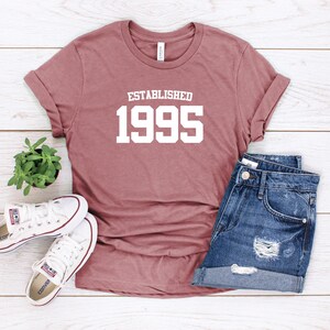 Puede incluir: Una camiseta color malva con el texto "ESTABLISHED 1995" impreso en blanco. La camiseta est&aacute; colocada sobre una superficie de madera blanca con un par de zapatillas blancas y unos pantalones cortos de mezclilla azul.