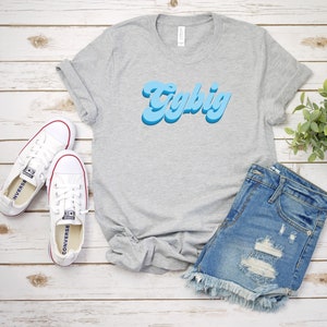 May include: A gray t-shirt with the text "Try Big" in blue retro lettering. The shirt is laid out on a white wooden surface with a pair of white canvas shoes and a pair of blue denim shorts.