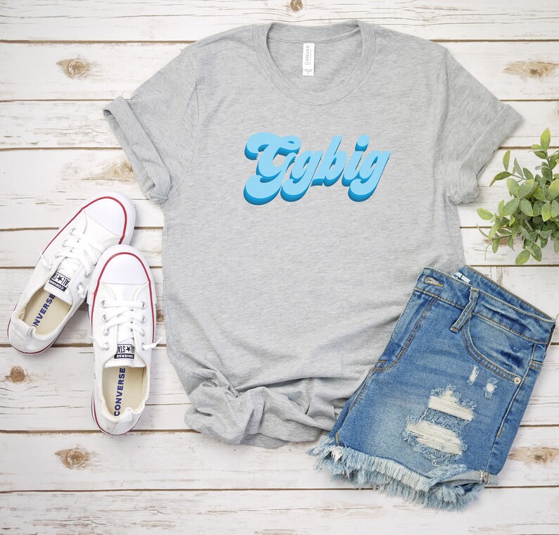 May include: A gray t-shirt with the text "Try Big" in blue retro lettering. The shirt is laid out on a white wooden surface with a pair of white canvas shoes and a pair of blue denim shorts.