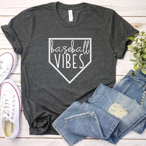 May include: A gray t-shirt with a white baseball diamond graphic and the text "baseball vibes". The shirt is laid out on a white wooden surface with a pair of white sneakers and blue jeans.