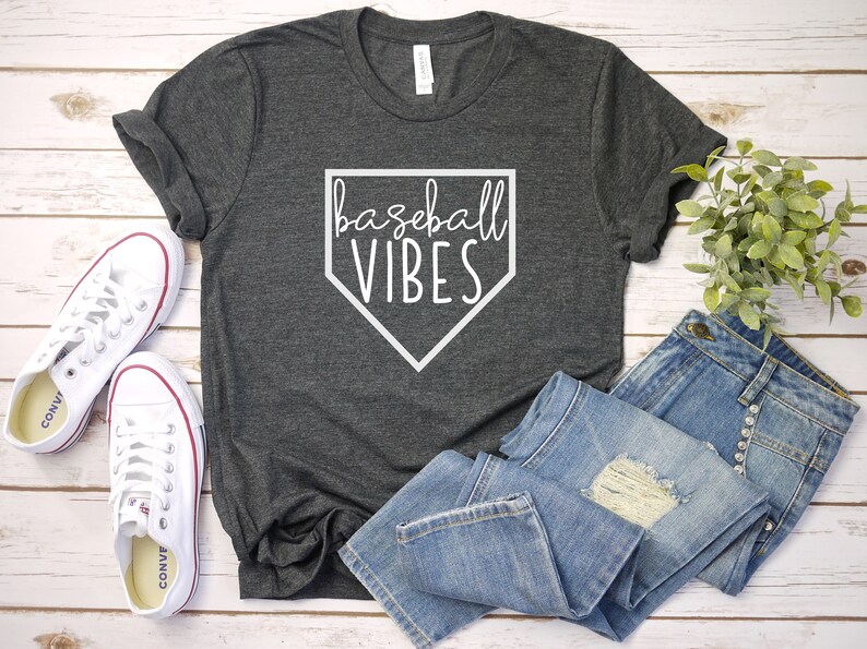 May include: A gray t-shirt with a white baseball diamond graphic and the text "baseball vibes". The shirt is laid out on a white wooden surface with a pair of white sneakers and blue jeans.