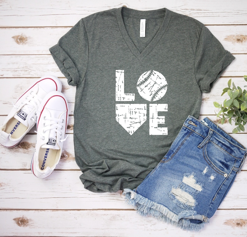 May include: A dark gray V-neck t-shirt with the word "LOVE" in white, featuring a baseball graphic. The shirt is paired with denim shorts and white sneakers with red accents, arranged on a light wood surface.