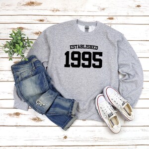 May include: Gray sweatshirt with black text that reads "Established 1995". The sweatshirt is paired with blue jeans and white sneakers.