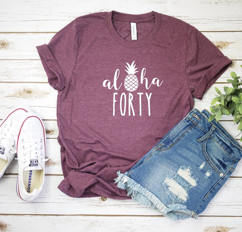 May include: A maroon t-shirt with the words "aloha FORTY" and a pineapple graphic in white. The shirt is paired with denim shorts and white canvas sneakers. The items are arranged on a light wood surface.