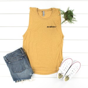May include: A mustard yellow sleeveless tank top with the word "mom" embroidered on the front. The tank top is laying on a white wooden surface with a pair of white sneakers and a pair of blue denim shorts.