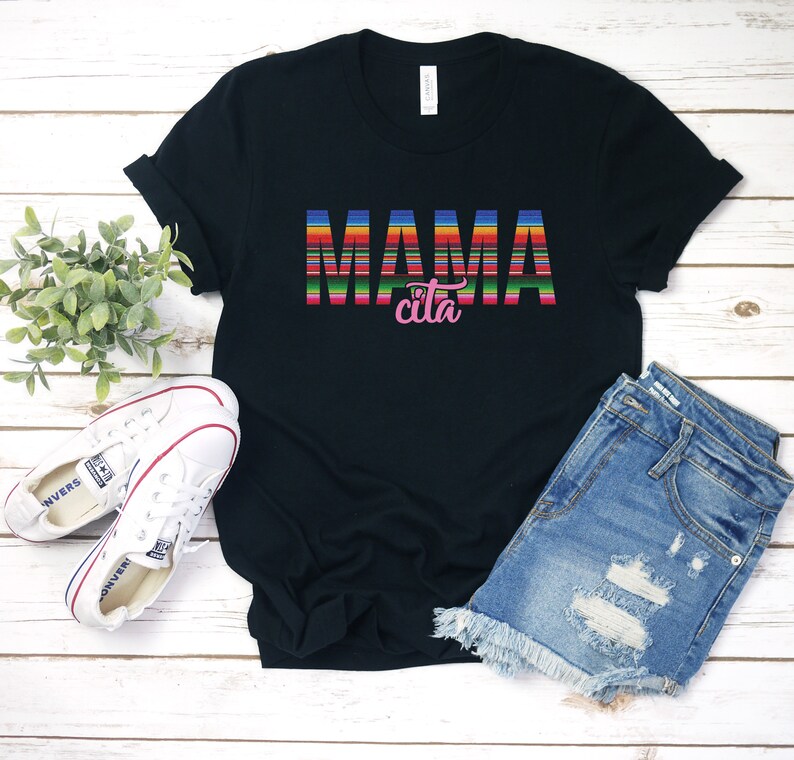 Op de afbeelding: Zwart T-shirt met de tekst "MAMA cita" in kleurrijke gestreepte letters. Het shirt ligt op een houten oppervlak met een paar witte sneakers en een blauwe denim short.