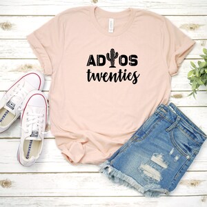 Puede incluir: Una camiseta rosa con el texto "ADIOS twenties" y un gr&aacute;fico de cactus. La camiseta est&aacute; sobre una superficie de madera blanca con un par de zapatillas blancas y unos pantalones cortos de mezclilla azul.