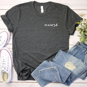 May include: A dark grey t-shirt with the word "nurse" embroidered on the front in white. The shirt is laid flat on a white wooden surface with a pair of white sneakers and a pair of blue jeans.