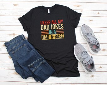 May include: Black t-shirt with a vintage-style graphic that reads "I keep all my dad jokes in a dad-a-base". The shirt is paired with blue jeans and gray sneakers.